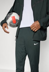 Grønn atletisk jakke med svarte detaljer, hvit t-skjorte, grønne bukser, Nike-logo. Person som holder en svart, hvit og rød mønstret fotball.
