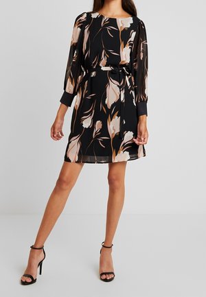 Frau trägt ein schwarzes Kleid mit floralem Muster, langen transparenten Ärmeln, einem Taillenband und schwarzen High Heels mit Knöchelriemen, steht auf einem hellgrauen Boden.