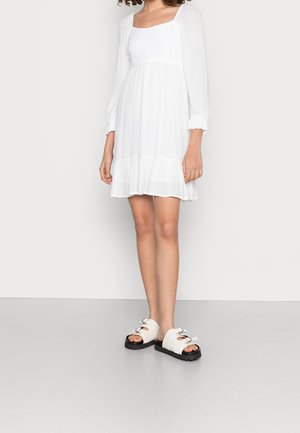 Mujer con vestido mini blanco de manga larga, con corpiño fruncido y falda de volantes, combinado con sandalias blancas de punta abierta y suelas negras.