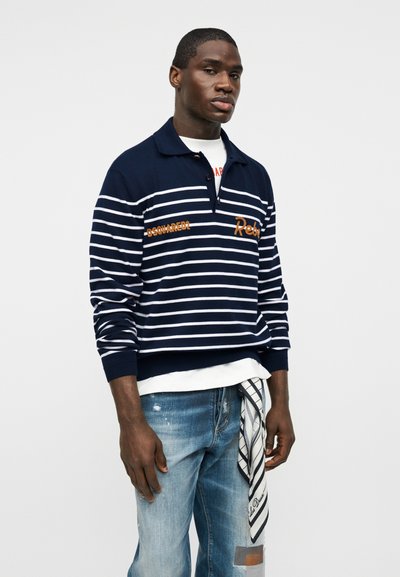 Homme portant un polo à manches longues rayé bleu marine et blanc, une chemise blanche en dessous, un jean clair usé avec une écharpe à motif accrochée à la poche.