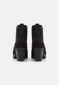 Paire de bottines en daim noir avec talons épais, panneaux latéraux élastiques et languettes à l'arrière, vue de dos sur fond blanc.