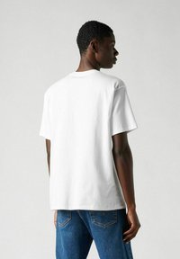 Camiseta de algodón blanca con mangas cortas y escote redondo, vista desde la espalda, combinada con jeans de mezclilla azul.