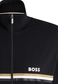 Schwarze Zip-Jacke mit hohem Kragen, weißen und goldenen horizontalen Streifen und einem Logo auf der linken Seite. Glatte Stoffstruktur.
