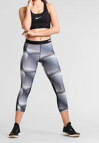 Czarny sportowy stanik i szare legginsy 3/4 w geometryczne wzory. Legginsy mają czarny pas w talii z logo "NIKE". Czarne buty sportowe.