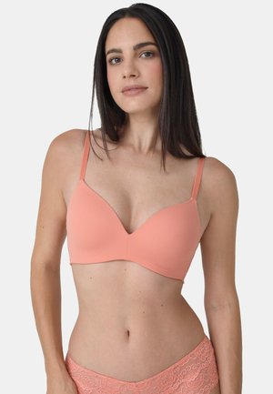 Frau mit langen dunklen Haaren, die einen korallenfarbenen BH und passende Spitzenunterwäsche trägt, vor einem schlichten hellen Hintergrund stehend.