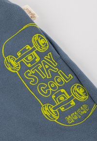 Blaue Baumwollbluse mit gelb bedrucktem Rücken, auf dem ein Skateboard-Design und der Spruch "STAY COOL" sowie der Markenname "sigikid" zu sehen sind.