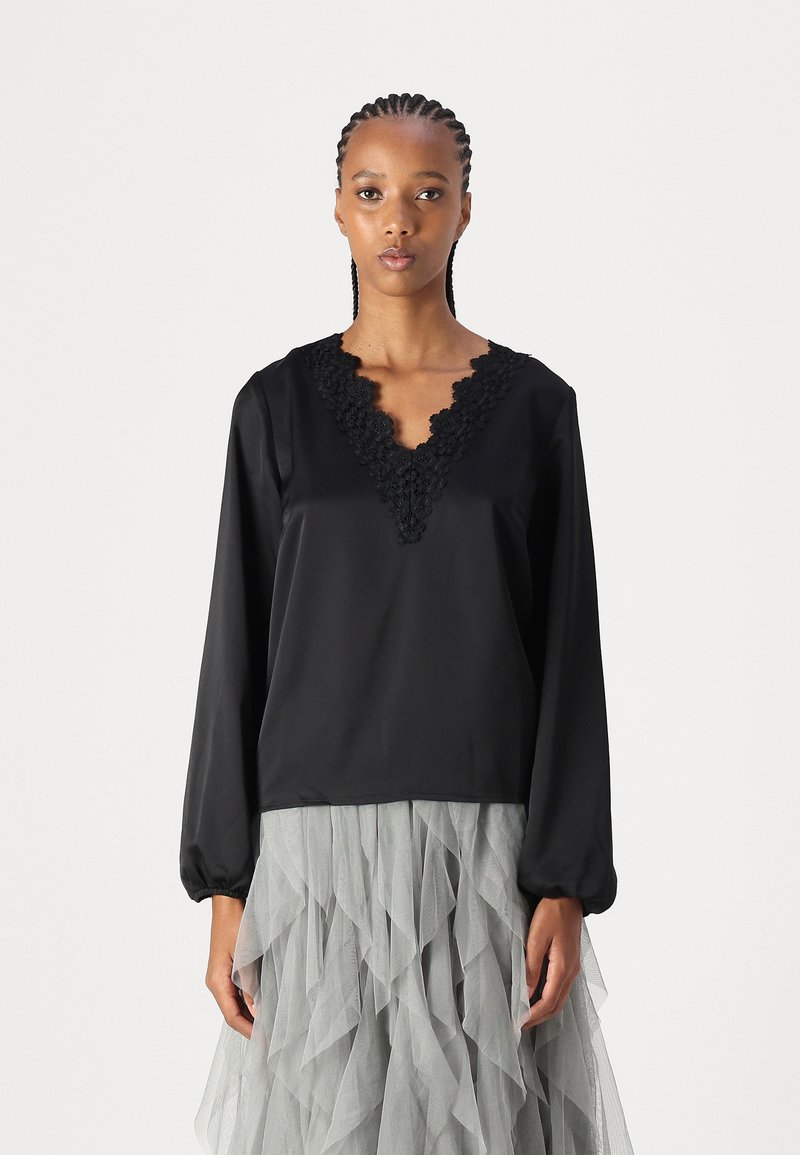 Blusa nera a maniche lunghe con scollo a V e dettagli in pizzo strutturato nel colletto. Tessuto di raso liscio. Abbinata a una gonna in tulle grigio a strati.