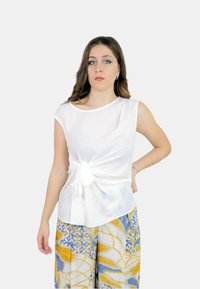 Top bianco senza maniche con dettaglio annodato sul davanti, abbinato a pantaloni larghi a fantasia con design floreali blu e gialli.