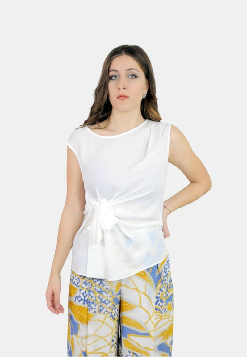 Top bianco senza maniche con dettaglio annodato sul davanti, abbinato a pantaloni larghi a fantasia con design floreali blu e gialli.