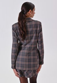 Grauer karierter Blazer mit offenem Kragen, strukturierten Schultern und einem dezenten Rüschensaum. Mit pinken und beigen Linien sowie einem rückseitigen Stickdetails.