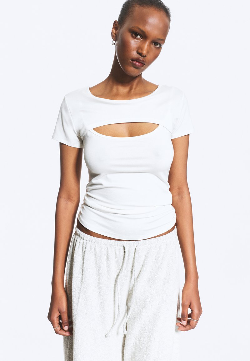 Femme à la peau foncée et aux cheveux tirés en arrière porte un haut blanc à manches courtes avec une découpe poitrine et un pantalon blanc ample avec cordon sur fond blanc.