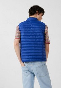 Persona joven vistiendo un chaleco acolchado azul brillante sobre una camisa de manga corta a rayas con vaqueros azul claro, de pie con las manos en los bolsillos traseros.