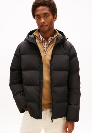 Chaqueta de plumas negra con capucha, con cierre de cremallera y costuras visibles, superpuesta a un suéter beige y una camisa de cuadros con cuello.
