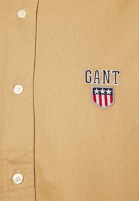 Lyhythihainen beige nappipaita, joka on valmistettu teksturoidusta kankaasta. Vasemmassa yläkulmassa on laivastonsininen kirjailtu logo, jossa on kilpi ja tähtiä.