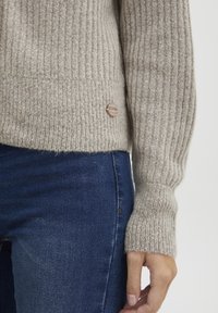 Lys beige ribbet genser med strukturert strikk, en tettsittende silhuett og en dekorativ knapp i kanten. Kombinert med mørkeblå jeans.