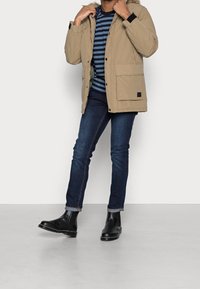 Muž upravující béžovou parku, oblečený v modro-proužkované košili, tmavě modrých džínách a černých kožen�ých Chelsea botách na šedé podlaze.