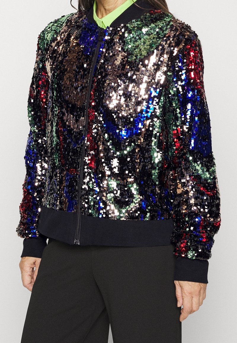 Femme portant une veste bomber multicolore à sequins par-dessus un top vert citron et un pantalon noir, debout devant un fond uni.