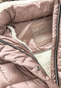 Interior de una chaqueta de plumón rosa con un suave forro de piel sintética, cremallera plateada y una etiqueta que presenta un nombre de marca y mensaje.