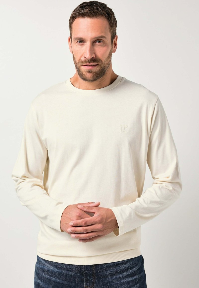 Cremefarvet langærmet t-shirt lavet af blødt stof, med en rund halsudskæring og et diskret præget logo på brystet, parret med mørke jeans.