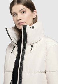 Veste en nylon léger beige clair avec un col haut, une fermeture éclair noire et des accents à cordon de serrage. Comprend des sections matelassées pour l'isolation.