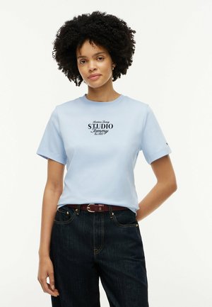 Femme aux cheveux bouclés portant un T-shirt bleu clair avec le texte "Studio Tommy" et un jean foncé, debout devant un fond blanc uni.