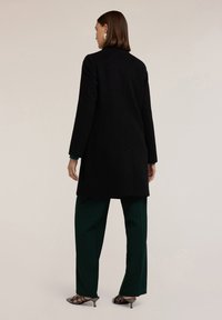 Cappotto in lana nera con forma dritta, chiusura a un bottone e vestibilità slim. Abbinato a pantaloni verdi e tacchi a punta neri.