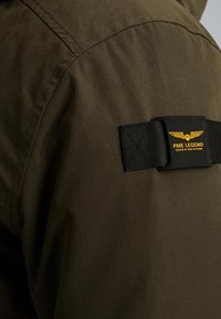 Szczegół oliwkowej kurtki z czarną naszywką z żółtym tekstem i logo skrzydeł, podkreślający trwały materiał i szycie.