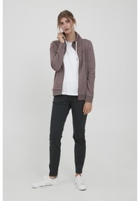 Mörkröd zip-hoodie med hög krage, ljusa snören, som är ihop med en vit skjorta och mörka slim-fit byxor. Vita sneakers fulländar looken.