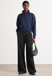 Navy pullover with a half-zip collar, ribbed cuffs, and hem. Paired with high-waisted black pleated trousers and a small black handbag.