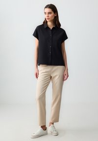 Camicetta nera a maniche corte con bottoni, dalla texture morbida, abbinata a pantaloni beige a gamba dritta e sneakers bianche. Design semplice e pulito.