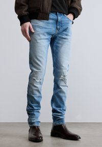 Homme portant un jean déchiré bleu clair, une chemise noire, une veste marron et des bottes en cuir marron foncé, debout sur un sol gris.