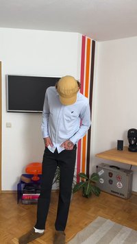 Lys blå langermet skjorte, svarte bukser, tan-kap, brune sko. Tatoveringer synlige på håndleddet. Innendørs setting med fargerik veggdekor.