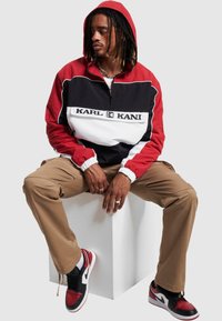 Punane, must ja valge poolsipullline kapuutsiga jakk, millel on logo; beežid püksid; mustad ja punased tossud valgete detailidega; tekstuuriga kangas.