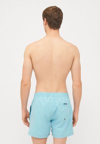 Pantaloni scurți de baie de culoare albastru deschis, cu un brâu elastic, o buzunare pe spate și o textură netedă. Lungime scurtă și design minim de cusătură laterală vizibil.