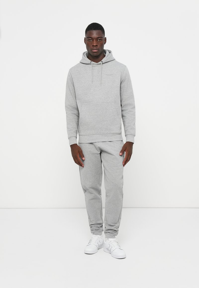 Sudadera de algodón gris con capucha y bolsillo tipo canguro, combinada con pantalones de chándal grises a juego, usados con zapatillas blancas. Diseño simple, ajuste relajado.