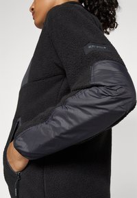 Zijaanzicht van een persoon in een zwart fleecejack met doorstikte nylon elleboogpatches en een ritszak voor de hand.