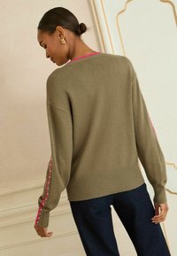 Olíva zöld, laza szabású kötött pulóver, rózsaszín díszítő varrással az ujjak mentén és bordázott szegéllyel. Sötétkék nadrág.