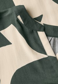 Tessuto verde e beige con un motivo, dotato di colletto e cuciture visibili. Superficie texturizzata, con forme astratte e colori a contrasto.