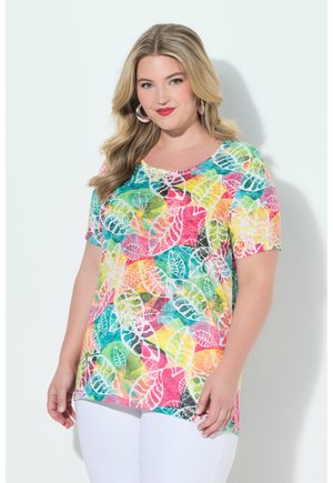 Frau trägt ein buntes, kurzärmeliges Oberteil mit Blattmuster und weiße Hose, hat lockeres blondes Haar und trägt Creolen, lächelt vor weißem Hintergrund.