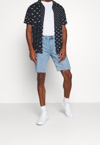 Kortärmad svart skjorta med knappar och vita geometriska mönster, i kombination med fransade ljusblå denimshorts och vita sneakers.