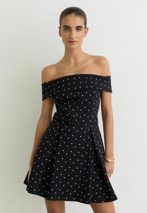 Kobieta w czarnej sukience z odkrytymi ramionami w białe kropki, z dopasowanym paskiem w talii, stojąca na tle jasnej, gładkiej ściany.