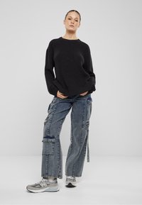 Schwarzer gerippter Pullover, weite hellblaue Cargo-Jeans mit Patches und graue Sneakers. Das Outfit hat eine lockere Passform und auffällige Nähte.