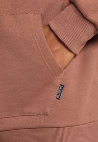 Felpa rosa con tasca a marsupio, polsini e orlo a coste. Presenta un'etichetta nera sul lato con il nome del marchio. Texture morbida.