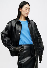 Giubbotto bomber in pelle nera con spalle imbottite, chiusura con zip e polsini a costine, indossato sopra un maglione azzurro chiaro e una gonna in pelle nera.