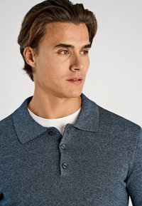 Koszulka polo z ciemnoniebieskiej dzianiny z usztywnionym kołnierzykiem, trzypanelowym klinem oraz gładką fakturą. Nosi się ją na białą podkoszulkę.