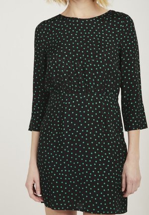 Vrouw die een zwarte jurk draagt met groene polkadots, driekwartmouwen en een getailleerde taille, gezien van nek tot midden bovenbeen.