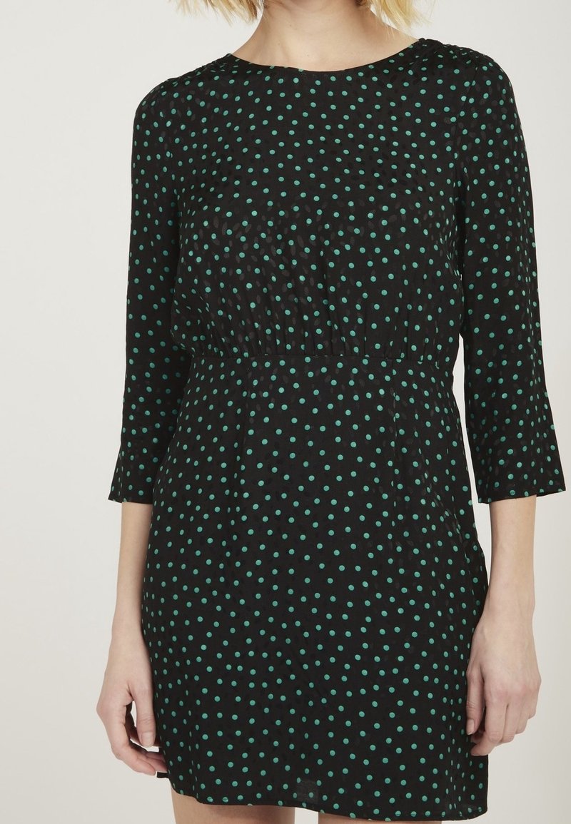 Femme portant une robe noire à pois verts, manches trois-quarts, taille cintrée, montrée du cou jusqu'à mi-cuisse.