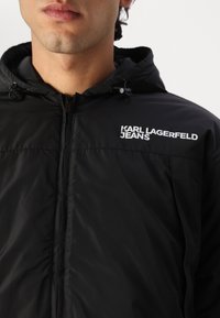 Black padded jacket with a hood, featuring a zippered front and a logo in white text. Smooth texture and fitted design.