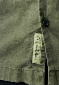 Olivově zelená košile vyrobená z texturované tkaniny, s černým knoflíkem a štítkem, na kterém je světle zeleným písmem napsáno "DURABLE GOODS LOT 002".
