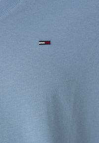 Svetlo modra bombažna majica s krožnim izrezom in majhnim vezenim logotipom, ki vključuje rdeče, bele in črne črte na prsih.